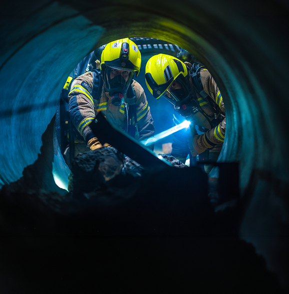 Rescue workers in a flooded sewer tunnel lift a stretcher through a sunlit overhead access opening.