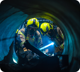 Rescue workers in a flooded sewer tunnel lift a stretcher through a sunlit overhead access opening.