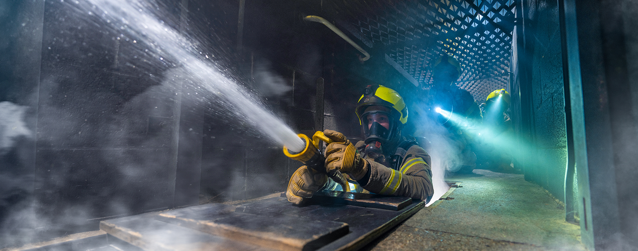 Firefighter sprays water through smoke-filled industrial corridor.